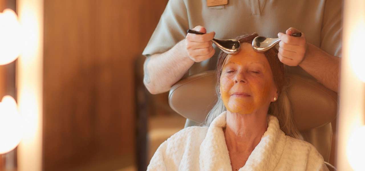 Client with an orange facial mask relaxes while a therapist massages her forehead using two metal spoons, seated in a spa chair, wearing a white robe under warm, soft lighting.