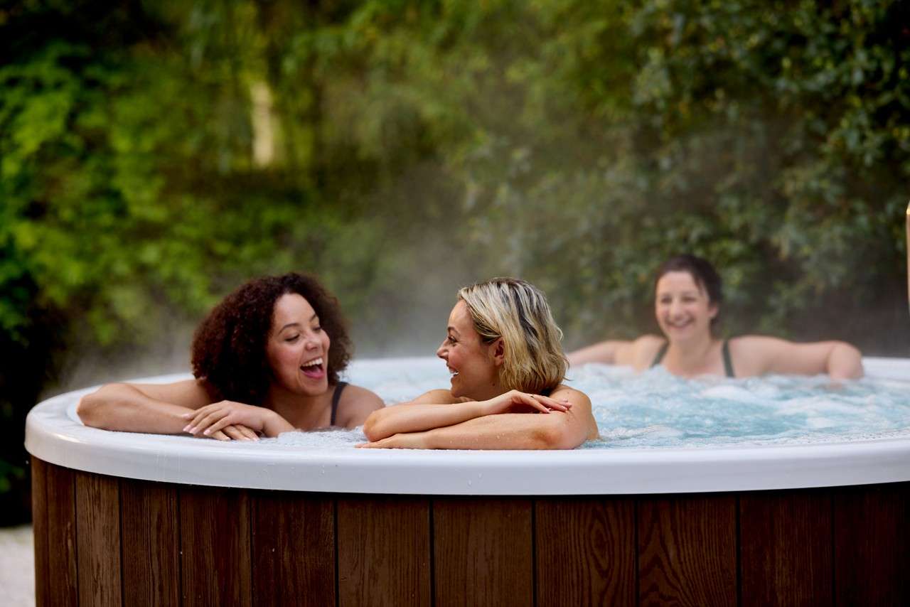 Three ladies in a warm bubbling hot tub. Two are holding on to the side looking at each other and laughing. 
