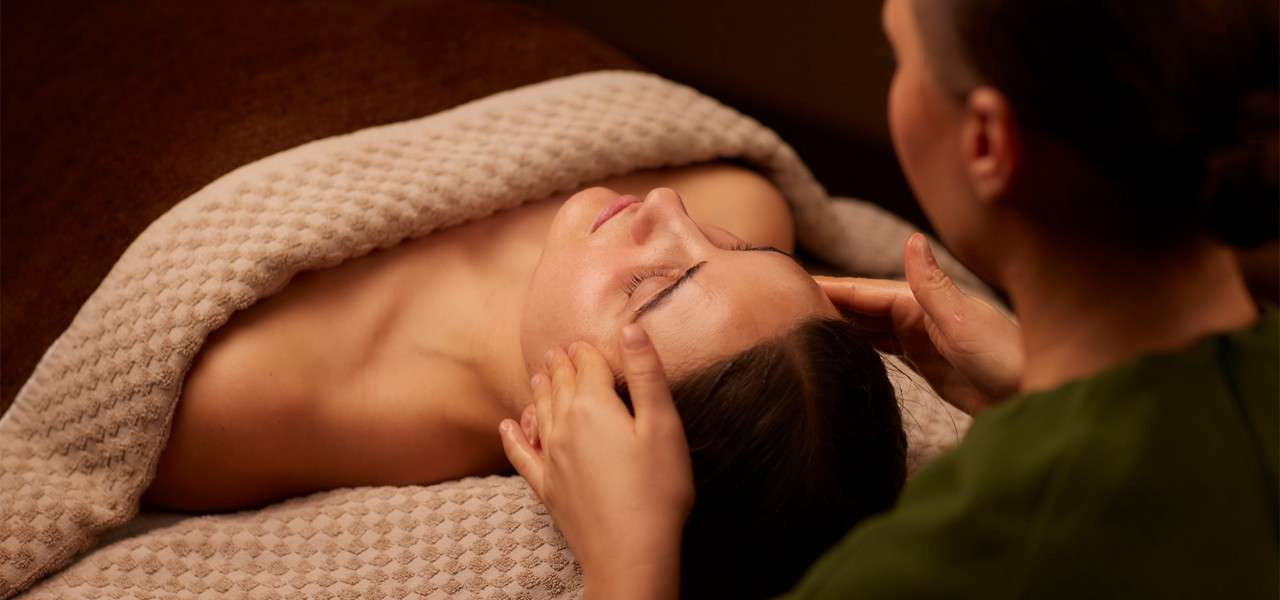 Client receives a gentle facial massage, eyes closed, while lying under a textured beige blanket; therapist’s hands cradle temples in a warm, softly lit spa room.