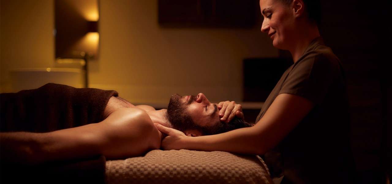 Massage therapist cradles a client’s head and neck, performing a treatment on a massage table in a dim, warm-lit spa room with towels and a softly glowing lamp.
