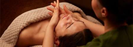 Person lies on a massage table; therapist gently presses their face with both hands; warm lighting and a textured blanket create a calm spa setting.