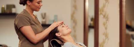Client reclines in a salon chair while a therapist gently massages their forehead, eyes closed. Mirrors, shelves, and soft lighting create a calm spa setting in the background.