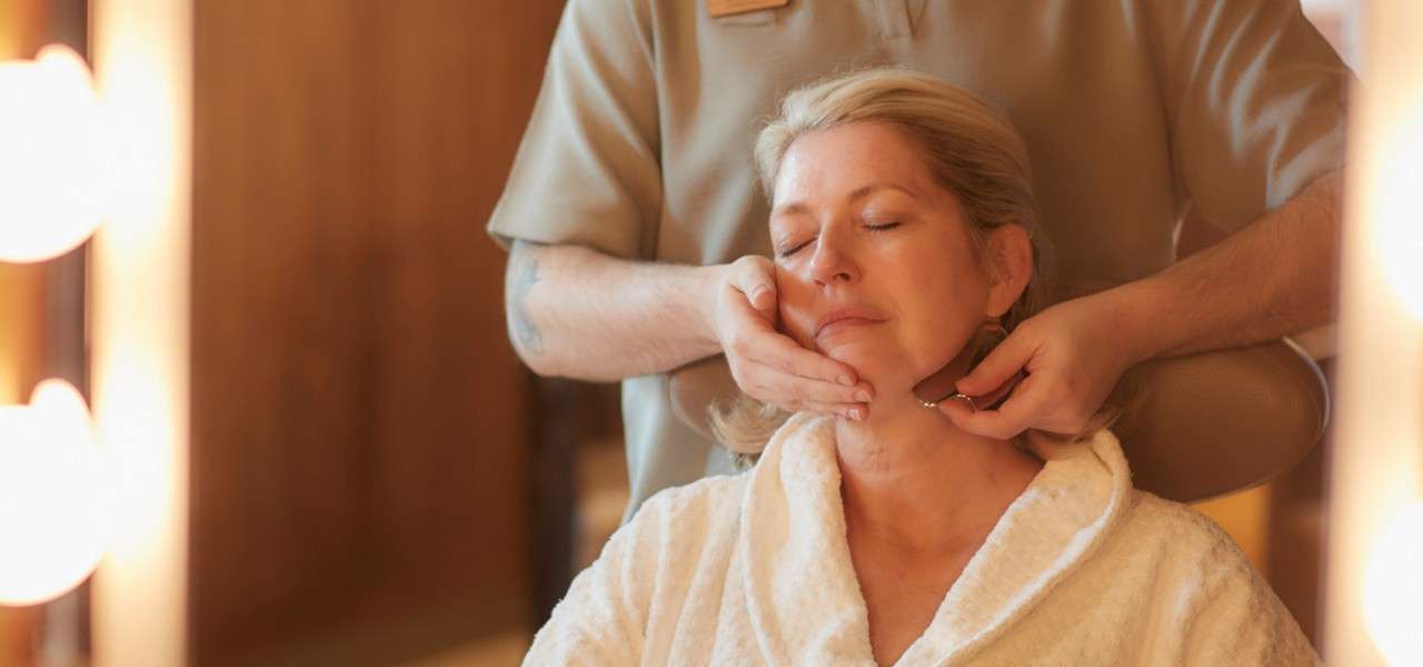 Client receives a facial treatment; therapist massages the chin with a small tool while she sits in a robe, eyes closed, in a warmly lit spa room.