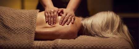 Person receiving a back massage lies face down on a table; therapist’s hands press and knead shoulders; soft lighting and a textured blanket create a calm spa setting.