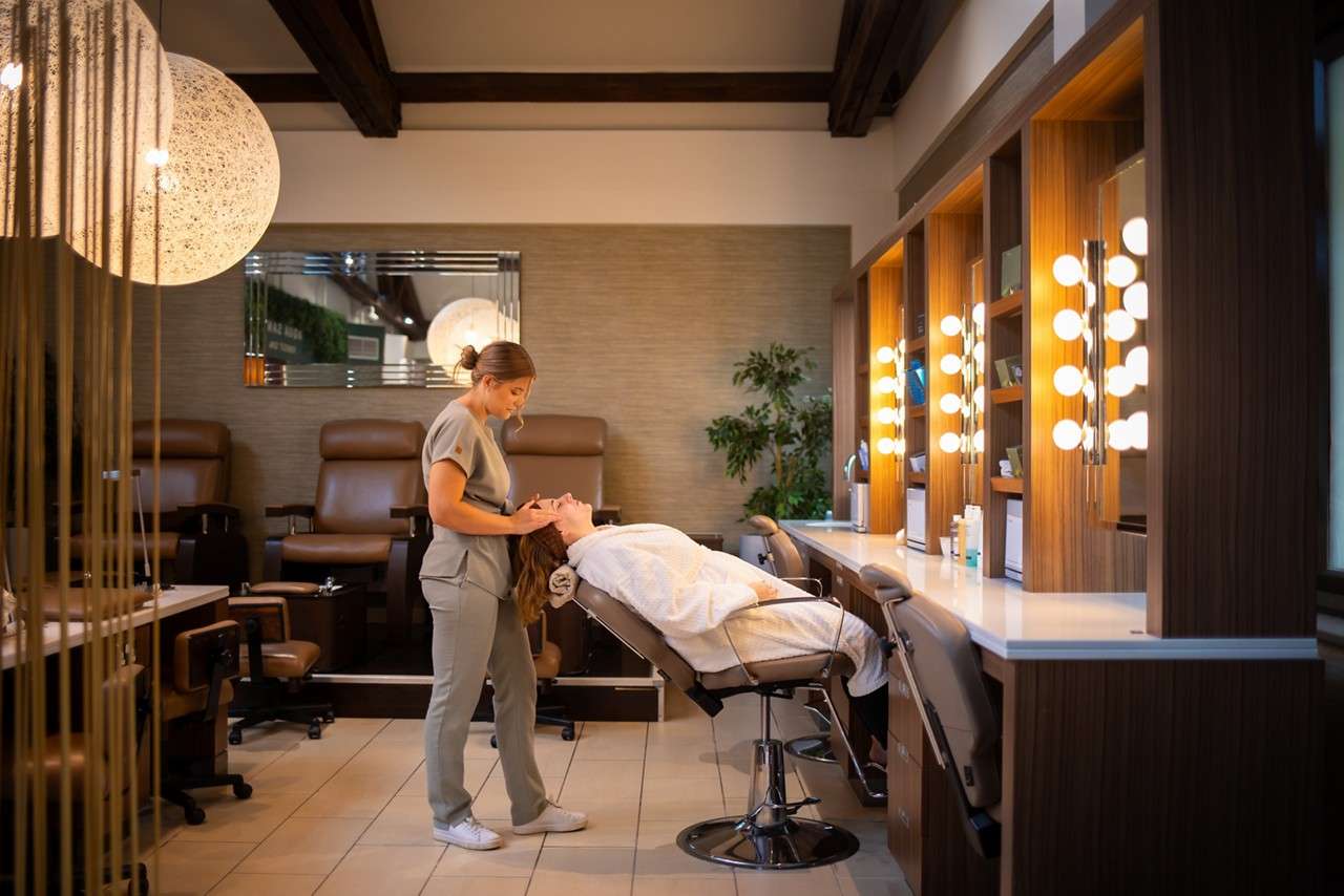 Esthetician massages a client's temples, the client reclining in a salon chair with headrest, eyes closed; warm-toned spa room with mirrors, shelves, and bottles in soft focus.