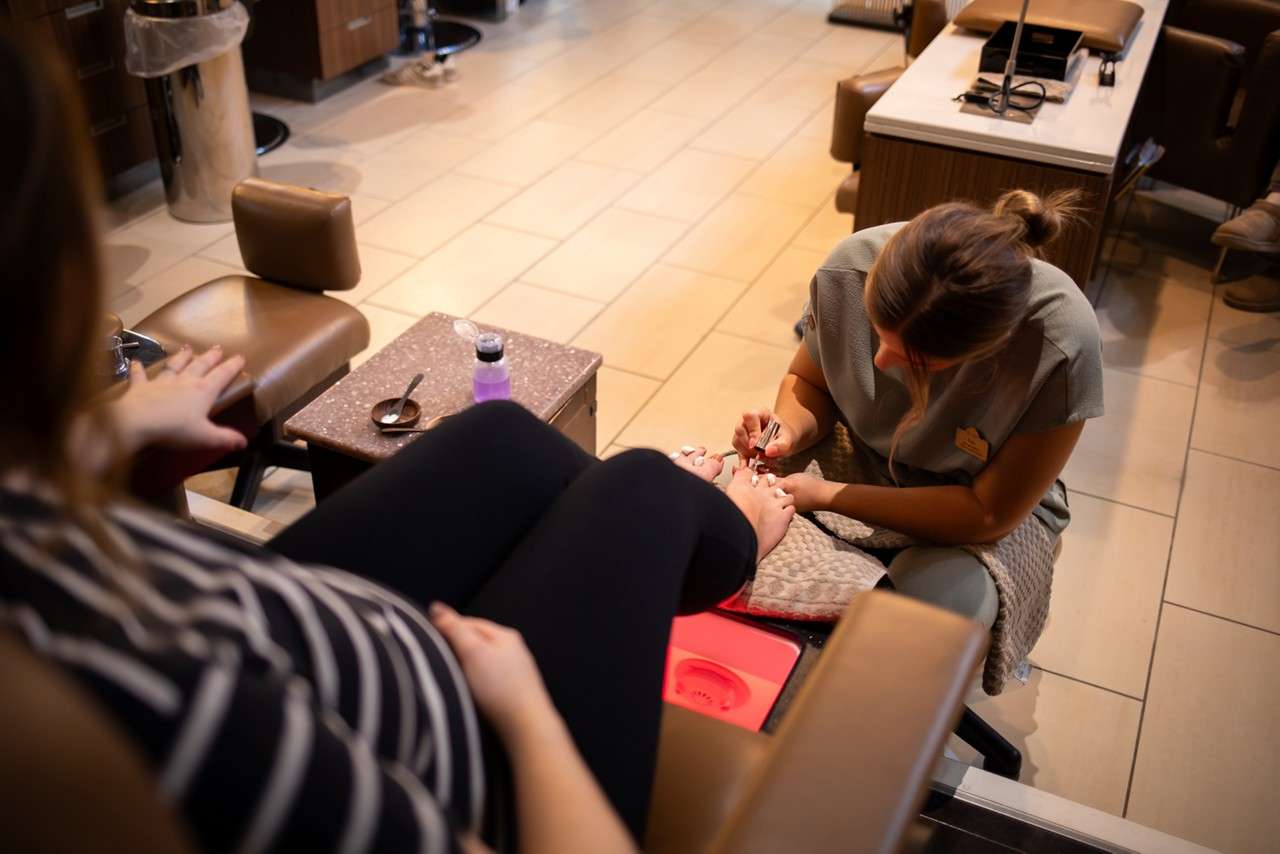Nail technician paints client's toenails beside a pedicure chair. Client reclines with feet above glowing foot bath; small table holds polish remover, tools, and towels in a salon.