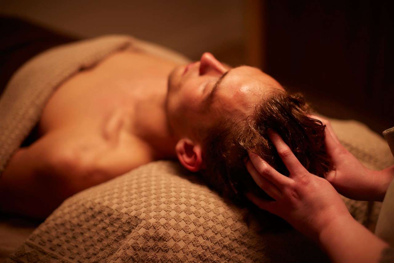 Client receives a scalp massage; hands press and knead the hairline as the person lies on a towel-covered table in a calm spa setting.
