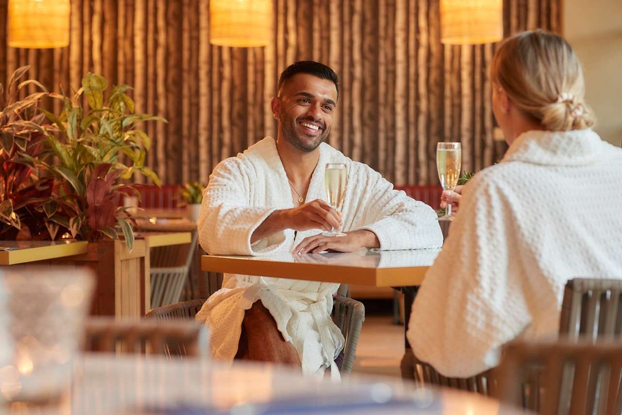 Couple in spa robes sat at a table with glasses of prosecco