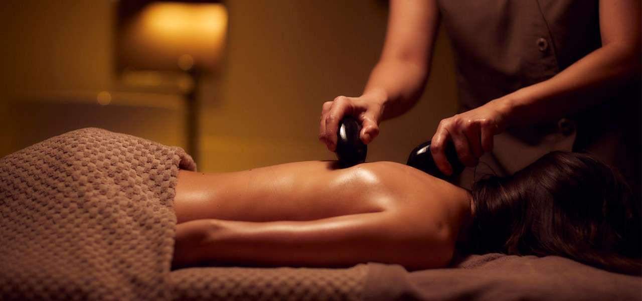 Client’s oiled back receives treatment; therapist presses and glides two hot stones along the spine; dimly lit spa room with warm lamp light, towel-covered massage table, tranquil atmosphere.