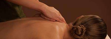 Person receiving a back massage; therapist’s hands press and slide along upper back and neck. In a spa setting with warm brown background; client’s hair tied in a bun.