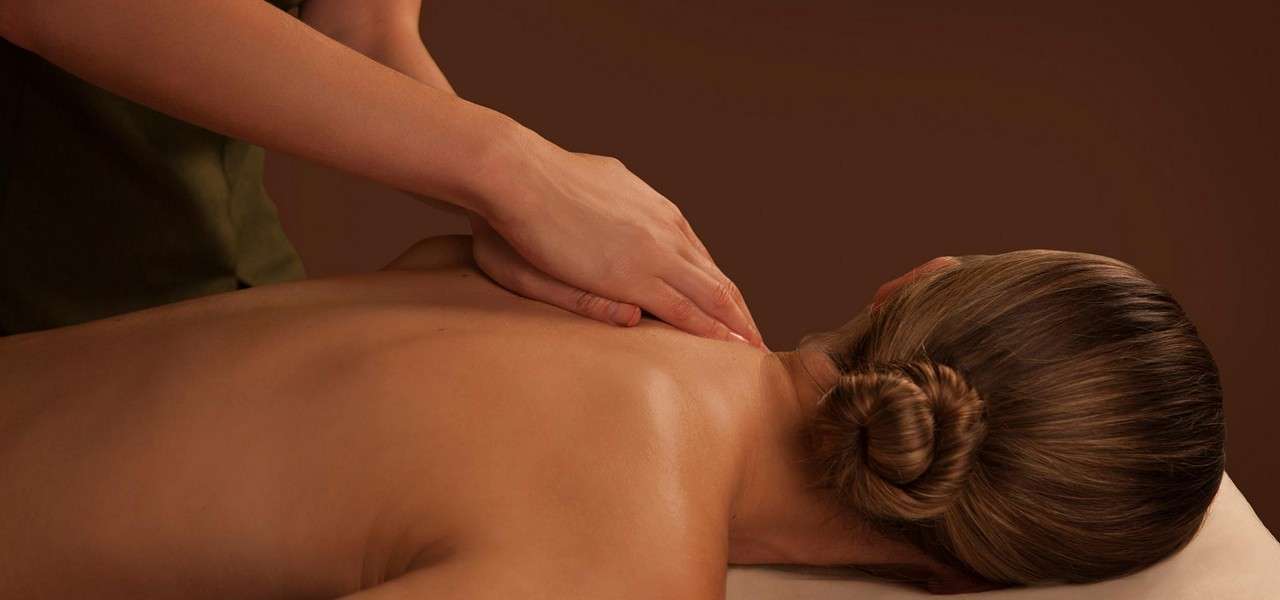 Hands massage a person’s upper back and shoulder, applying pressure. The person lies face-down on a massage table, hair in a bun, under warm, softly lit spa surroundings.