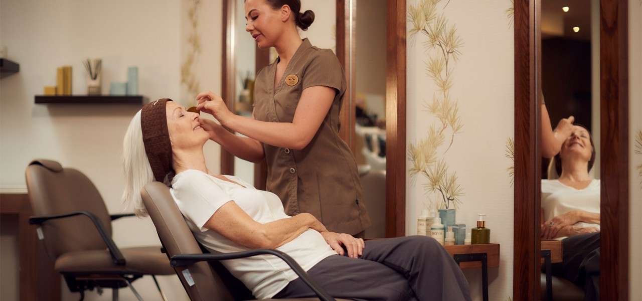 Beautician applies makeup to a reclining older woman in a salon chair; mirrors reflect them, and shelves hold products and towels. Visible text: VOYA.