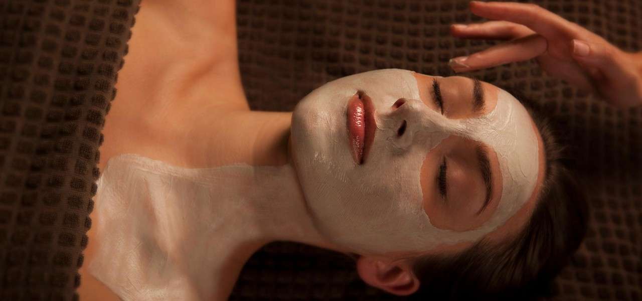 Person with a white facial-and-neck mask lies with eyes closed, relaxing; an attendant’s hand hovers near the forehead in a dimly lit spa, atop a textured brown blanket.