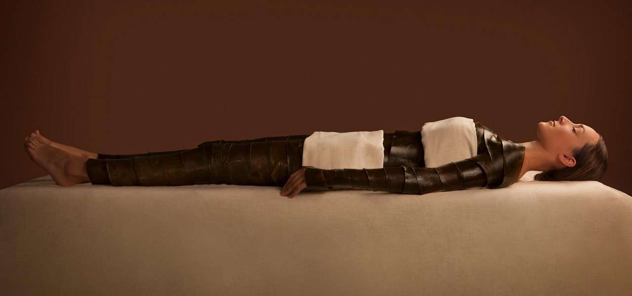 Person lies supine, wrapped in dark strips and white towels, on a padded table, in a spa-like setting with a warm brown background.