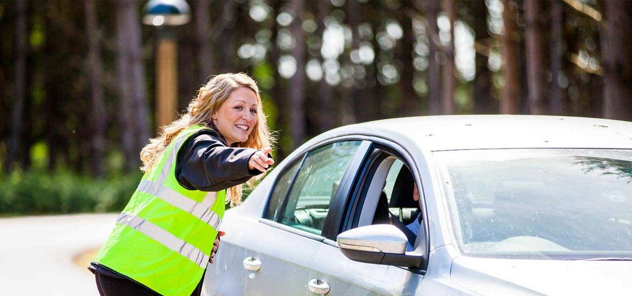 Traffic marshal in a fluorescent safety vest leans toward a silver sedan, pointing and smiling. She directs the driver at a roadside area bordered by tall, blurred trees.