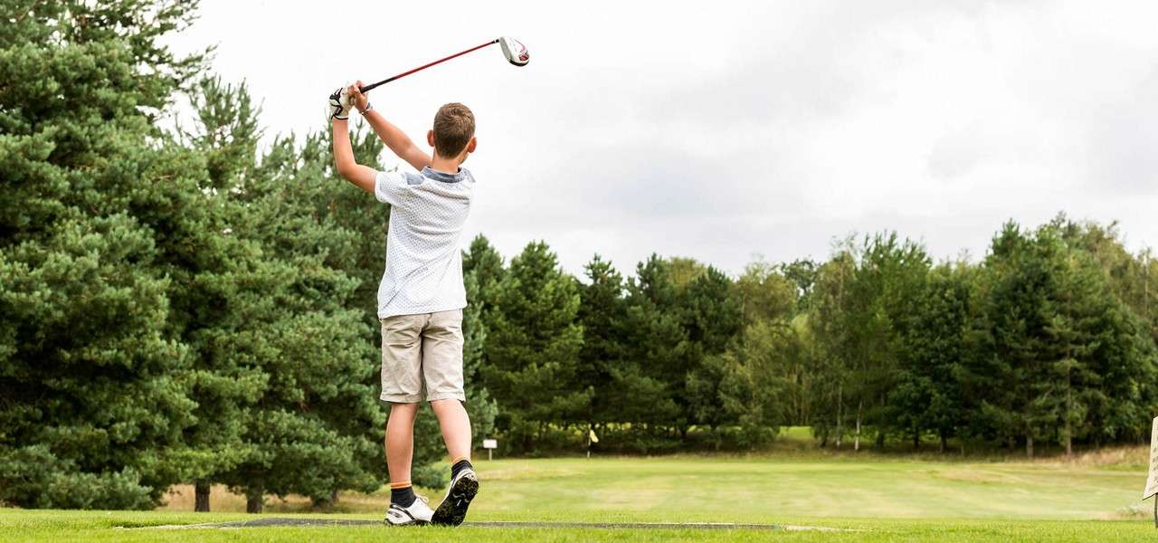 Young golfer swings a driver, finishing the follow-through on a tee box; open fairway stretches ahead, bordered by dense trees under an overcast, cloudy sky.