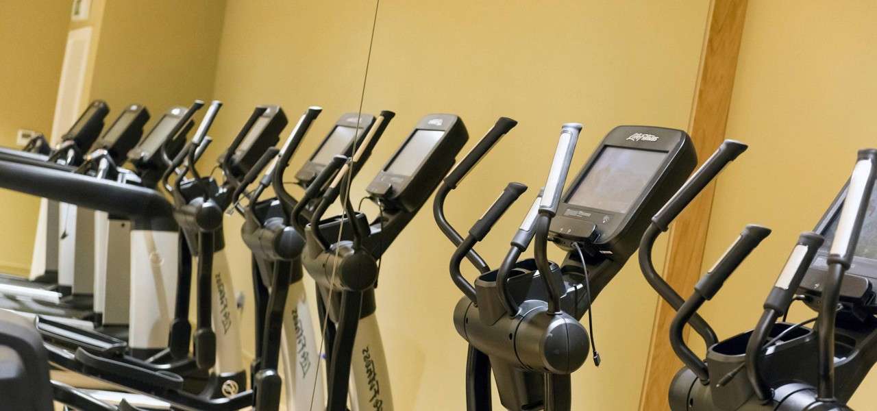 Elliptical machines stand idle, their handles and consoles aligned, in a quiet gym; a mirror reflects the row against beige walls. Text: Life Fitness.