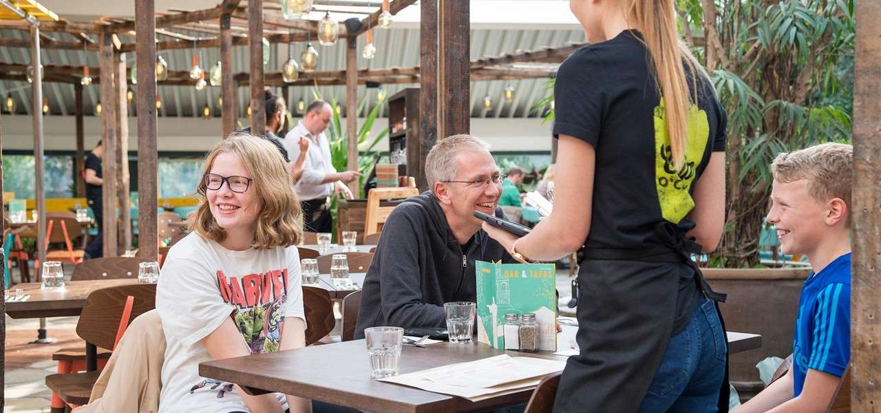 Server takes order from three smiling diners at a wooden-table restaurant. Hanging lights and plants surround. Visible text: "MARVEL" on a shirt; "BAR & TAPAS" on a menu.