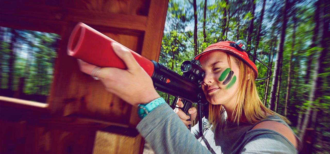 Person with face paint aims a scoped laser-tag rifle, squinting through the sight. They wear a red helmet and wristwatch, standing in a wooden shelter amid a sunlit forest.