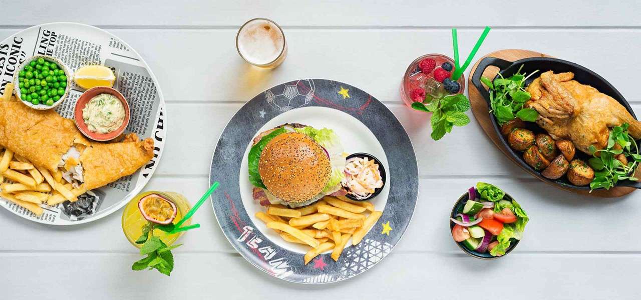 Plated meals sit on a light table: fish and chips with peas and tartar; a burger with fries and slaw; roast chicken with potatoes; side salad and drinks. Visible text: TEAM; newspaper-style print with unreadable words.