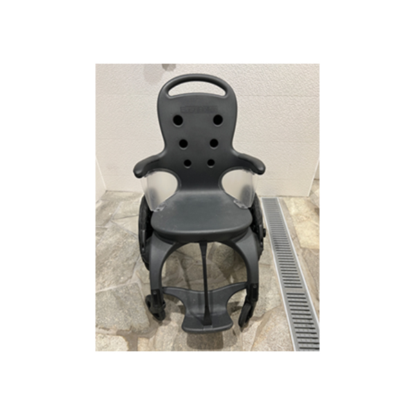 Child bicycle seat rests upright, displaying armrests, ventilation holes, and footrests with straps, in a tiled room beside a linear floor drain. Text on headrest: unreadable raised brand mark.