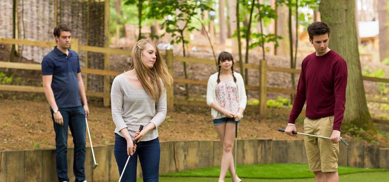 Four young adults hold putters, pausing on a mini-golf green. Two stand foreground preparing to putt; two watch behind. Tall trees, wooden fence, and shaded forest surround the course.