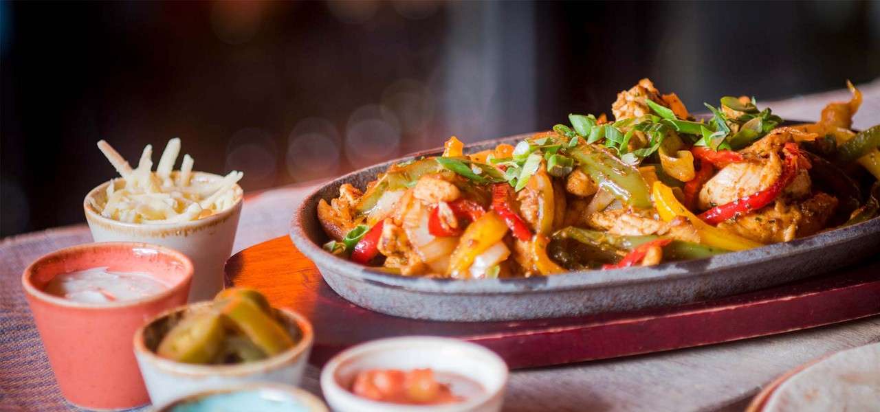 Fajitas sizzle in a cast-iron skillet, chicken, peppers, and onions glistening, while small bowls of cheese, jalapeños, salsa, and sauces surround it on a wooden board.