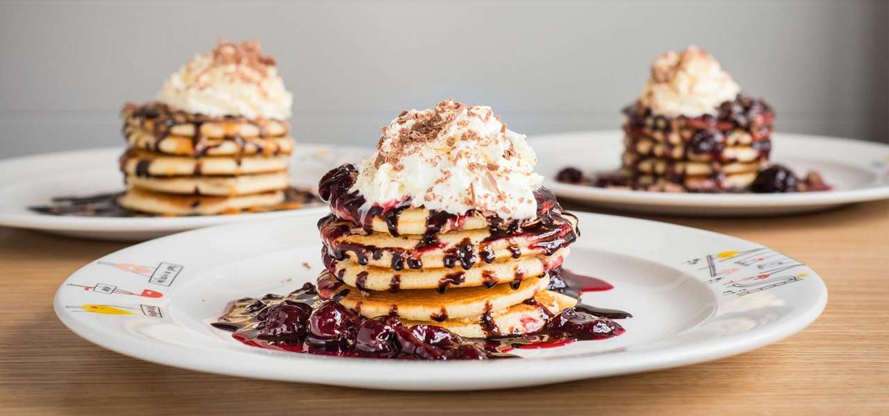 Stacked pancakes drip berry syrup, crowned with whipped cream and chocolate shavings; on a white patterned plate at a wooden table, with two similar pancake stacks softly blurred in the background.