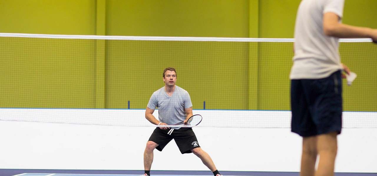 Man with badminton racket crouches ready position, watching opponent. Another player at right prepares to serve a shuttlecock across a net on an indoor court with lime-green walls.
