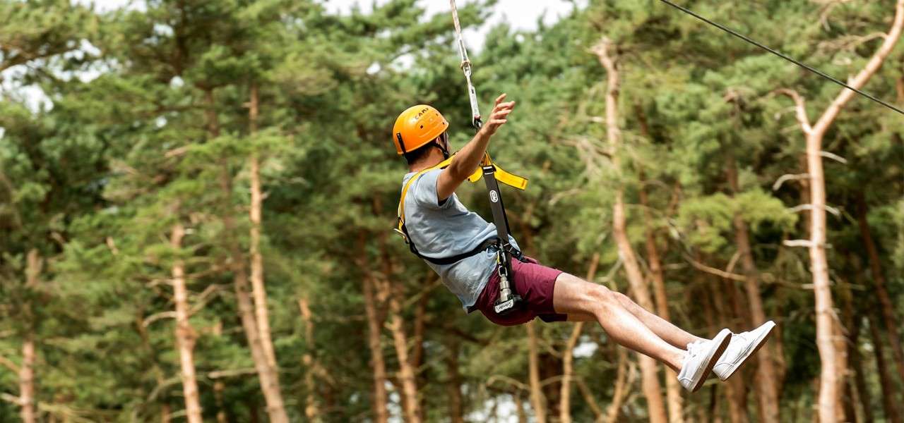 Person zip-lining, leaning back with one arm raised, legs extended, wearing harness and orange helmet. Forest trees fill the background. Text: “CAMP” on helmet.