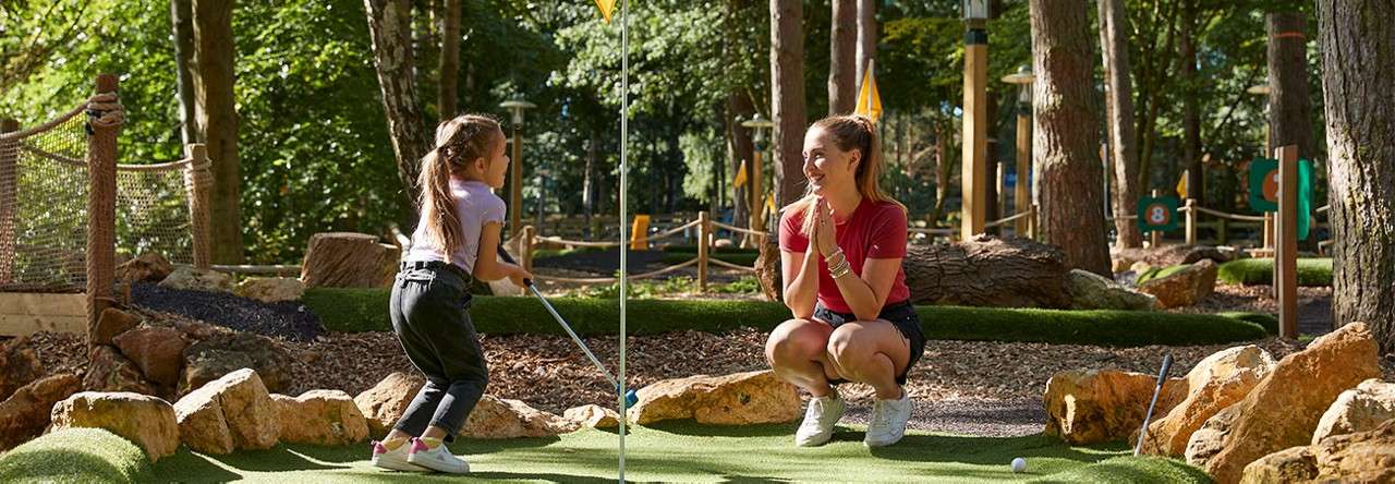 Child golfer swings a putter while an adult crouches, clapping encouragement; both on an artificial green beside rocks and flagsticks, within a sunlit, wooded mini-golf course.