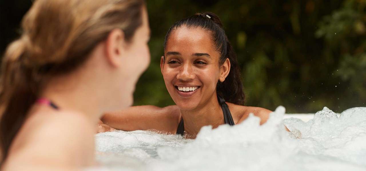 Two people soak and chat, smiling, in a bubbling hot tub. Steam and froth surround them. Background shows soft-focus greenery, suggesting an outdoor setting and relaxed, social atmosphere.