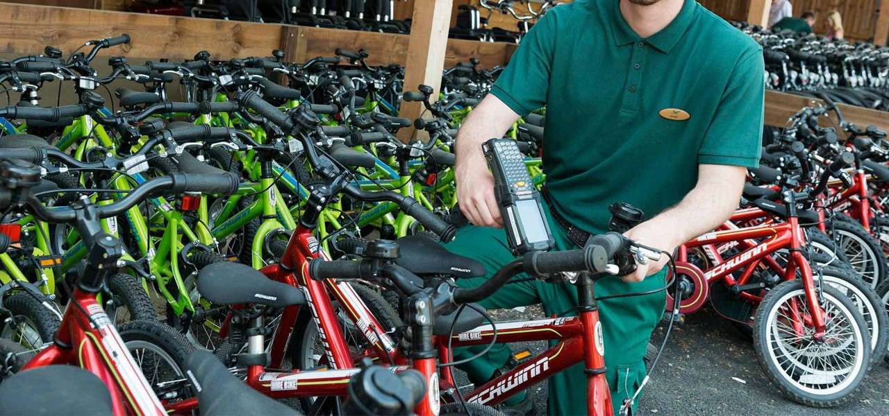 Worker scans a red children’s bicycle with a handheld device, amid rows of red and green bikes in an outdoor rental storage area. Text: SCHWINN; HIRE BIKE.
