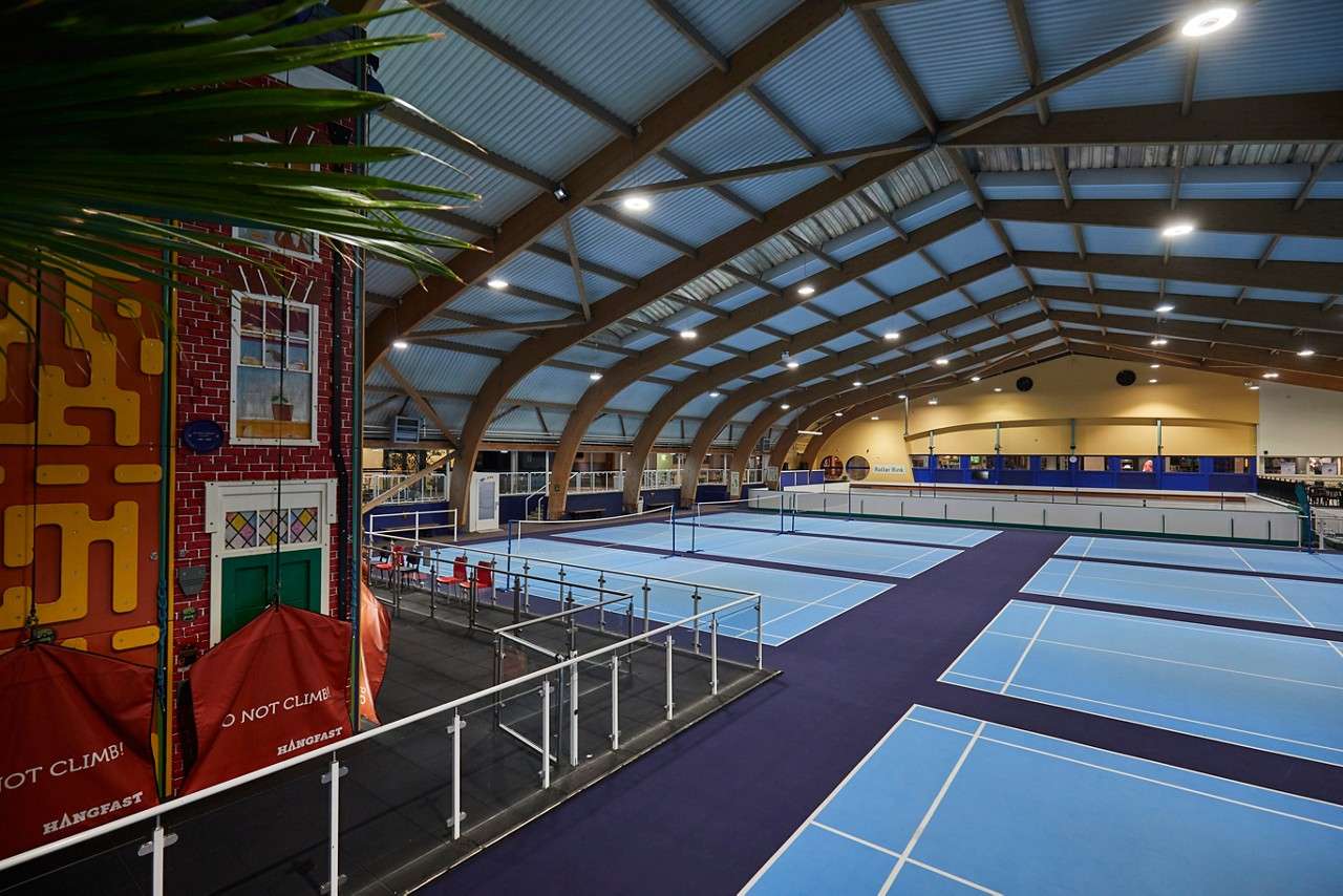 Badminton player crouches ready with racket, facing an opponent. Indoors on a bright green-walled court, a net divides them as he anticipates the next shot.