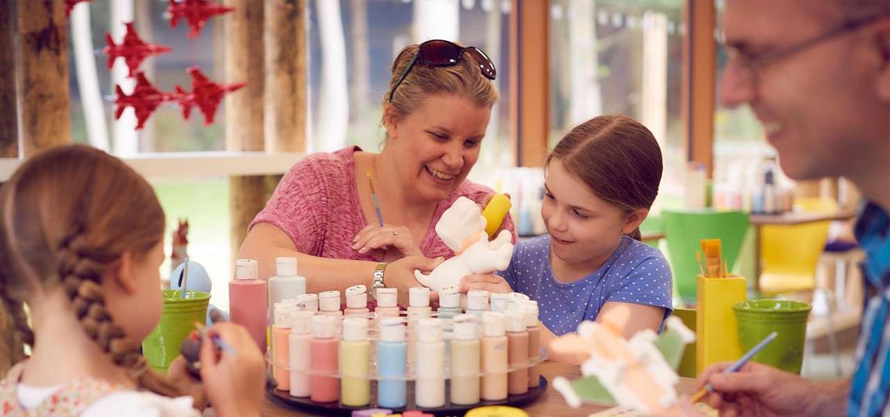 Family paints ceramic figurines, children and adults brushing colors onto a bunny. Context: bright studio with spinning rack of pastel paint bottles, windows, green chairs, and hanging red star decorations.