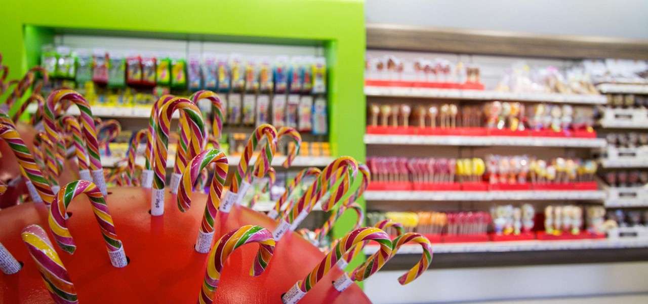 Candy canes stand upright in a red display, their rainbow stripes curving outward, while behind them shelves of lollipops and packaged sweets line a brightly lit, green-accented candy shop.