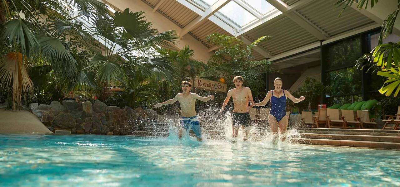 Three swimmers run forward, splashing into a turquoise pool; around them, palms, rock edging, lounge chairs, and skylights define an indoor tropical waterpark. Visible sign: “VENTURE BAY”.