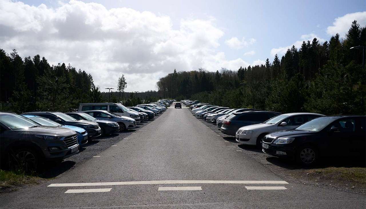 Cars line both sides of a wide asphalt lane, parked facing inward; a single vehicle drives away toward a distant forest under a bright, cloudy sky.