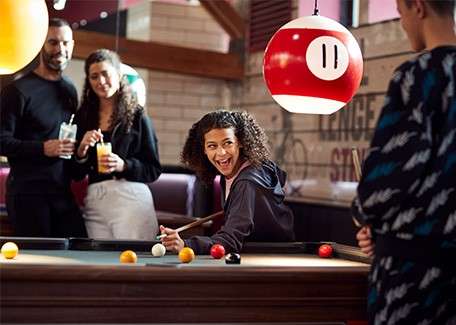 Young woman aims a pool cue, smiling; friends watch nearby in a casual bar with brick walls and a hanging billiard-ball lamp labeled “11”.