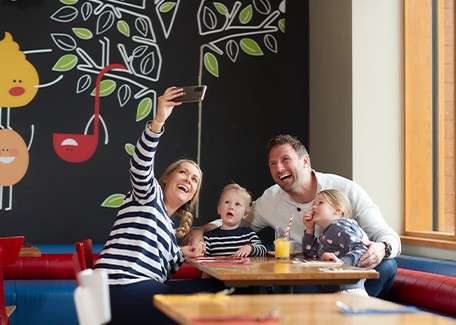 Family of four takes a selfie, smiling together at a cafe table; two children sit between parents with juice, beside a colorful mural wall and a large window.