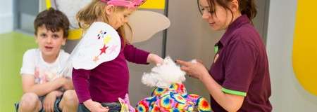 Girl wearing fairy wings interacts with a stuffed animal held by an adult, while a boy watches nearby; scene set in a bright indoor play or activity area.