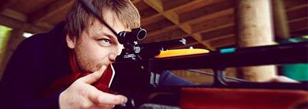 Person aims a scoped crossbow, pressing cheek to the stock and sighting forward; context: covered wooden shooting range with blurred background, suggesting focus and motion toward a distant target.