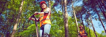 Two people ride Segways, smiling and leaning forward, wearing helmets, vests, and pads, moving along a forest path surrounded by tall trees and sunlight filtering through green leaves.