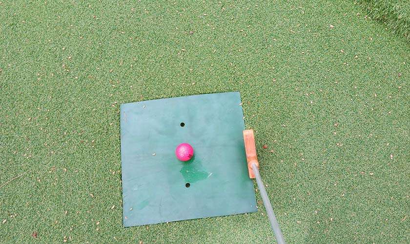 Pink mini-golf ball waits to be struck by a wooden putter, positioned on a small green square mat, set on artificial turf scattered with tiny debris.