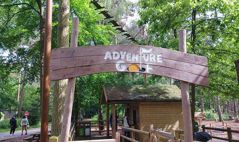 Wooden entrance sign announces: ADVENTURE GOLF. Beneath it, a small hut serves the course. People stroll nearby as a rope-bridge course runs overhead among tall, leafy forest trees.