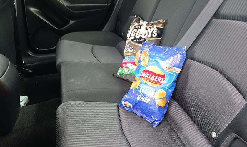 Two multipack crisp bags rest on a car’s rear seat; text visible: WALKERS CHEESE & ONION 6 PACK; McCoy’s CLASSICS.