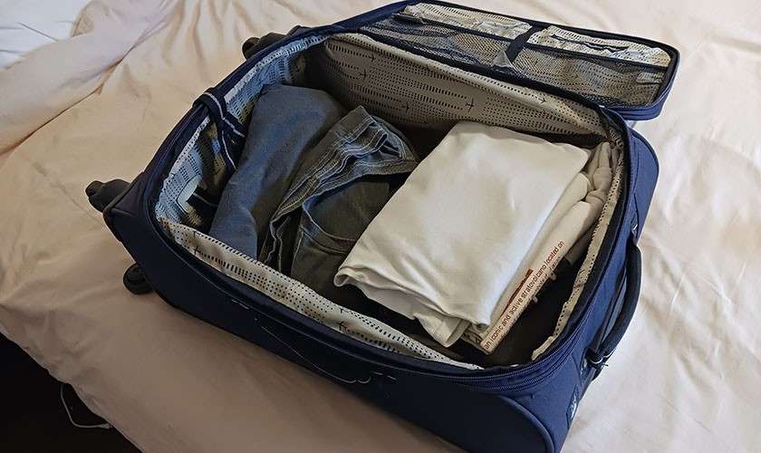 Suitcase opens, displaying folded clothes—white T-shirts and blue jeans—arranged inside; mesh zip compartments and lining visible. Context: resting on a bed in a softly lit room.