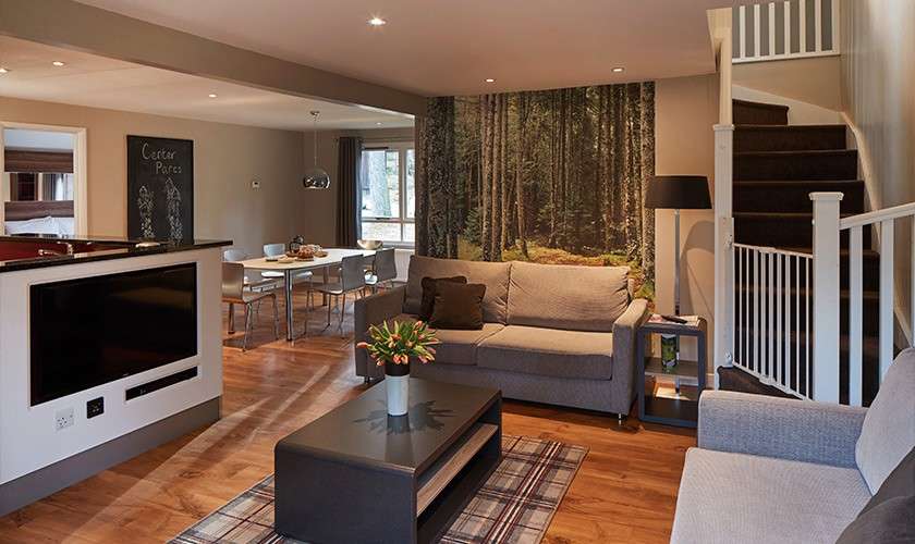 Sofa set frames a low coffee table with flowers, facing a wall-mounted TV; nearby, a staircase ascends and a forest mural decorates the wall; dining area beyond. Text: “Center Parcs”.