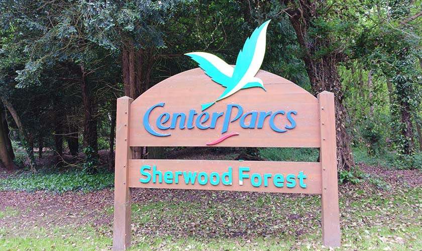 Wooden entrance sign displays “Center Parcs” and “Sherwood Forest,” featuring a stylized green bird logo, standing amid leafy trees and ground cover at a forest clearing.