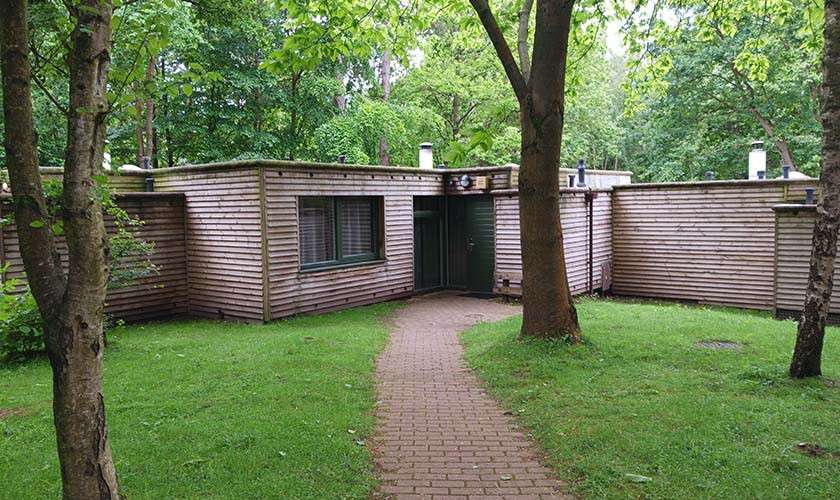 Single-story wooden cabin sits among trees; a brick path curves toward its central dark doorway. Rectangular windows with blinds face the grassy lawn, surrounded by dense green foliage.